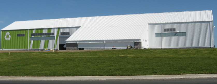 Materials rcovery facility. Large white building with light green stripes on the left. On the far left is a white recycling symbol in white. Blue sky background around the building with a green lawn in the front.